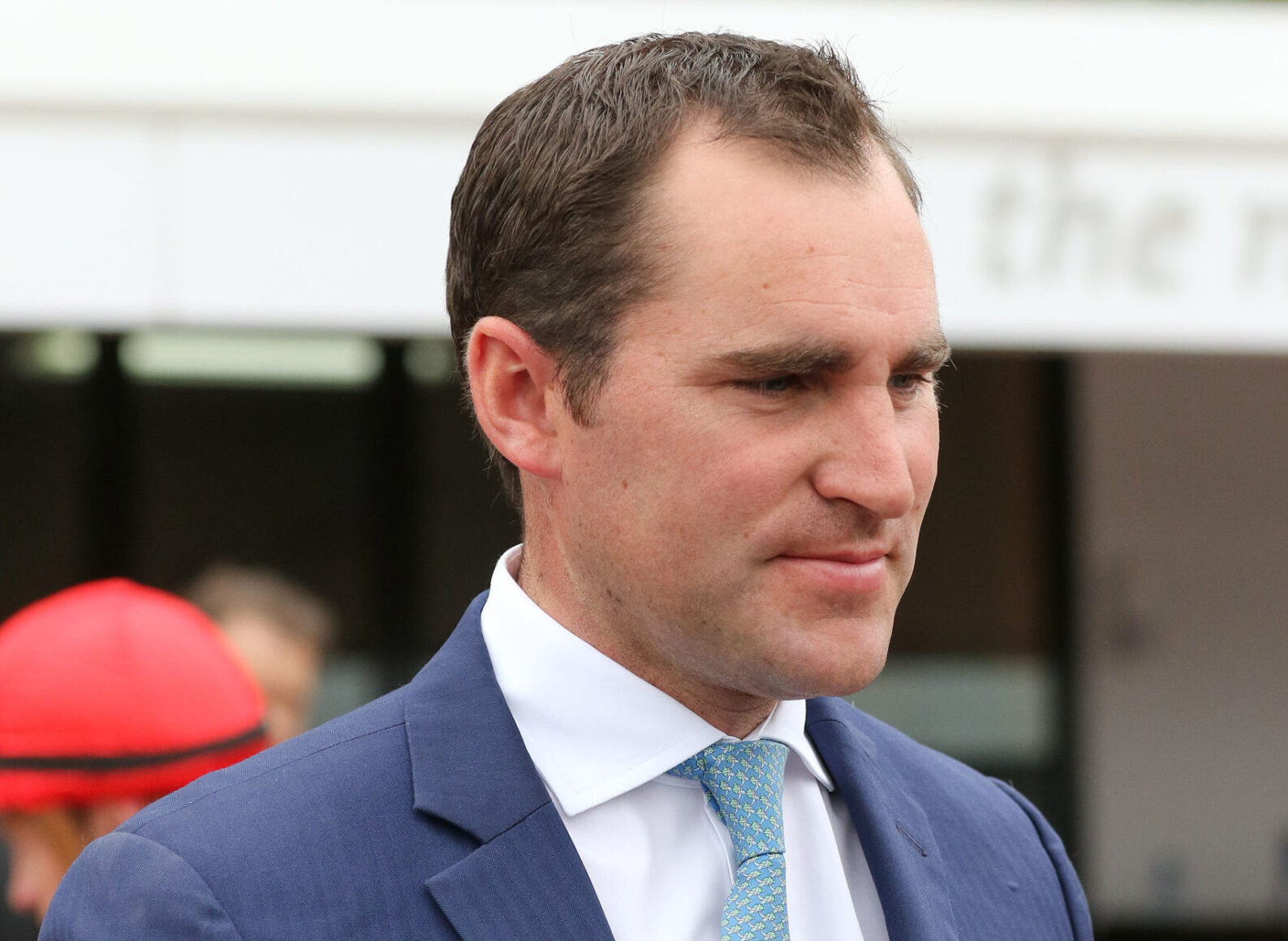 Man in a navy suit with a light blue patterned tie, looking to the right at an outdoor event or venue.