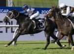Two jockeys sprint on racing horses on a grassy track, vying for the lead near the railing.