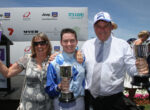 Three people celebrate on stage with trophies at a race awards ceremony, sponsor banners in the background under a blue sky.
