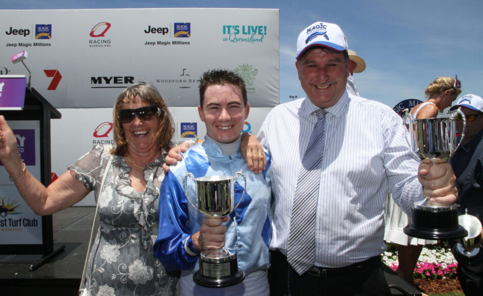 Three people celebrate on stage with trophies at a race awards ceremony, sponsor banners in the background under a blue sky.