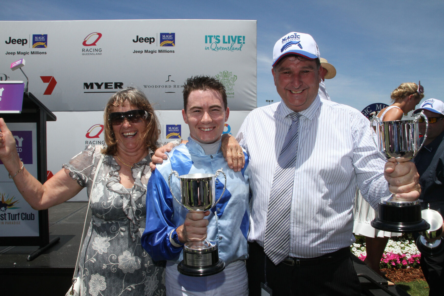Three people celebrate on stage with trophies at a race awards ceremony, sponsor banners in the background under a blue sky.