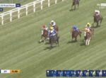 Group of thoroughbred horses with jockeys in colorful silks racing on a grassy track, white rails on the side, during a live Royal Ascot broadcast with on-screen race timers and odds at the bottom.