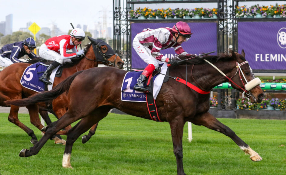 A brown horse leads a sprint at Flemington as jockeys push for the finish on a grassy racecourse, colorful banners in the background.