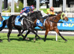 Two racehorses and jockeys sprint along the track, close to the inside rail, wearing blue/red and green/orange silks.