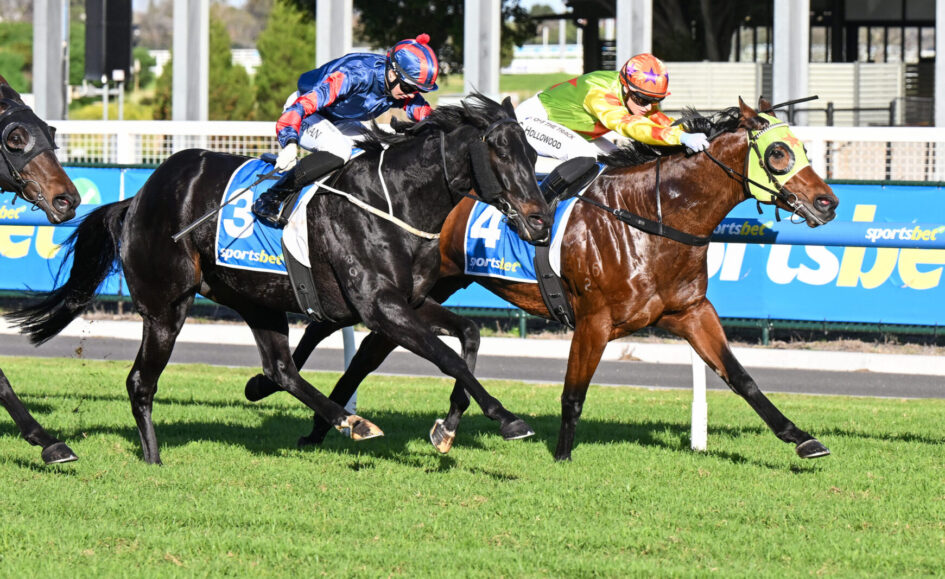 Two racehorses and jockeys sprint along the track, close to the inside rail, wearing blue/red and green/orange silks.