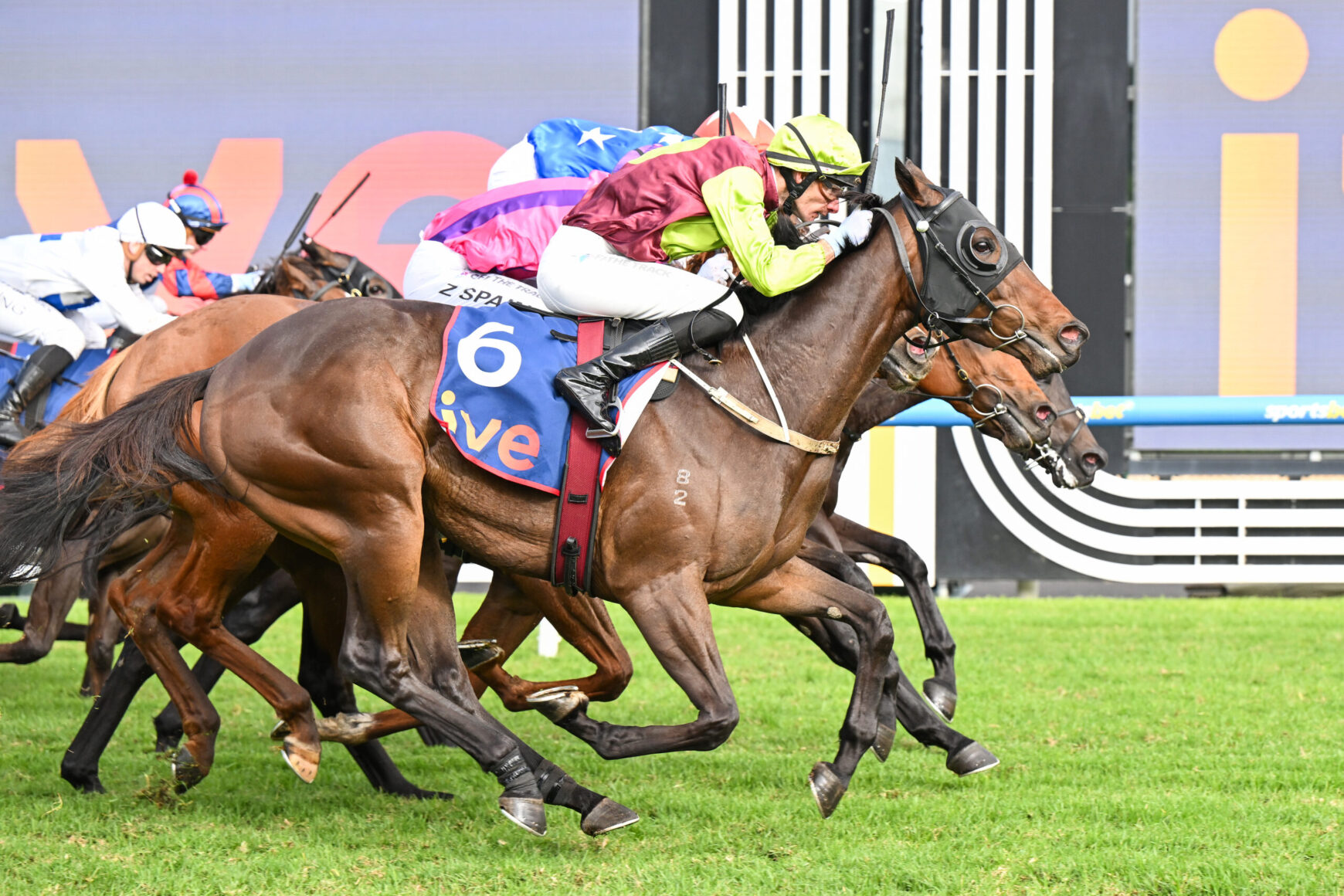 Close-up of a horse race in progress: a brown horse with a neon-lime and maroon-clad jockey leads, others close behind on colorful silks.