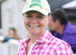 Smiling woman wearing a beige cap with 'Magic Millions' and a pink checkered shirt outdoors.