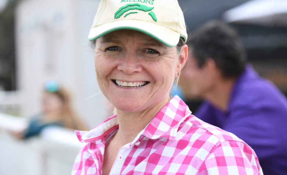 Smiling woman wearing a beige cap with 'Magic Millions' and a pink checkered shirt outdoors.