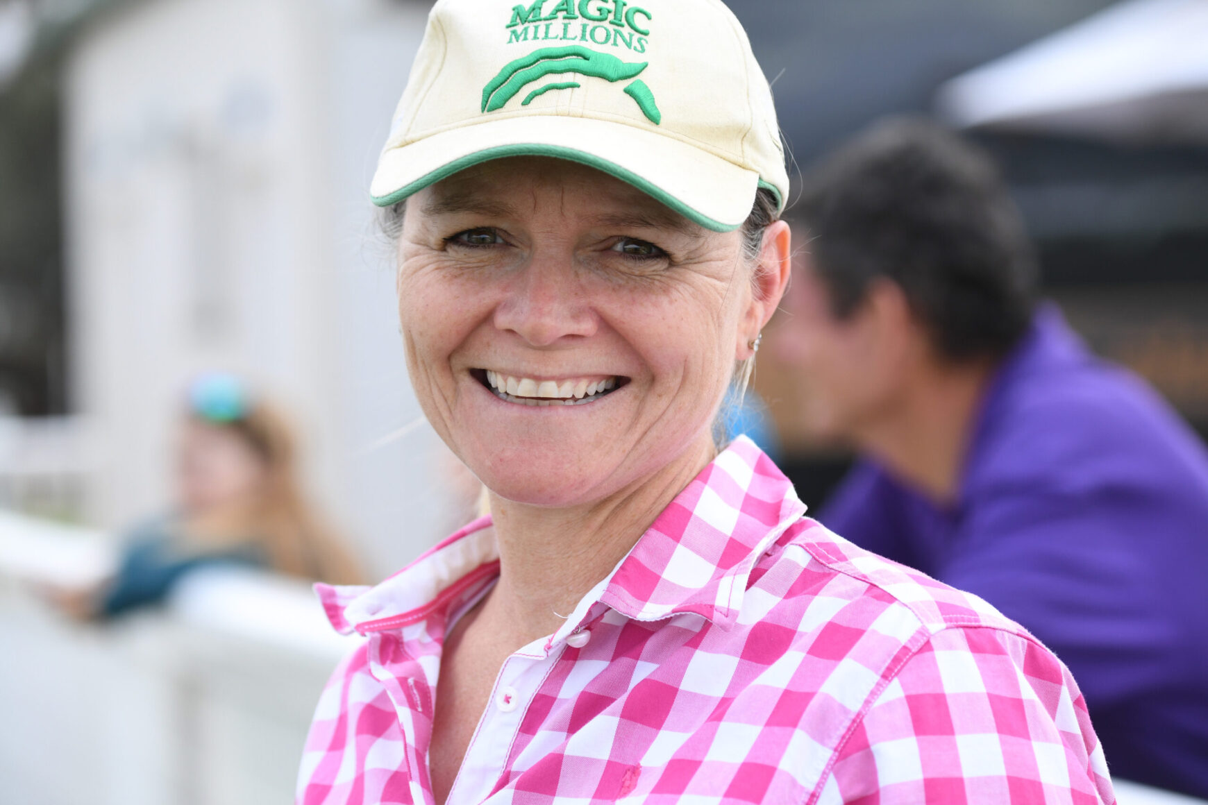 Smiling woman wearing a beige cap with 'Magic Millions' and a pink checkered shirt outdoors.