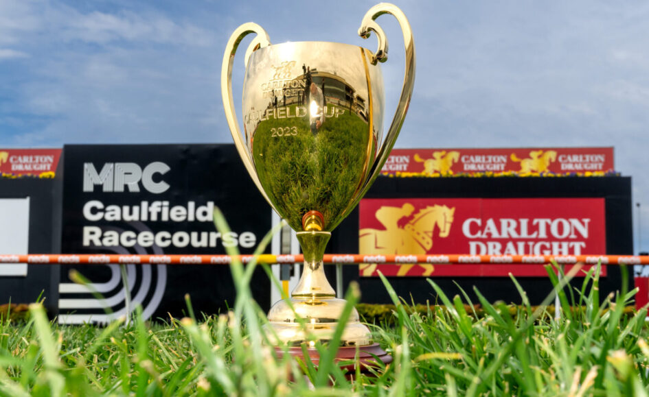 Gold trophy engraved with Carlton Draught Caulfield Cup 2023, standing in grass with racecourse signage in the background.