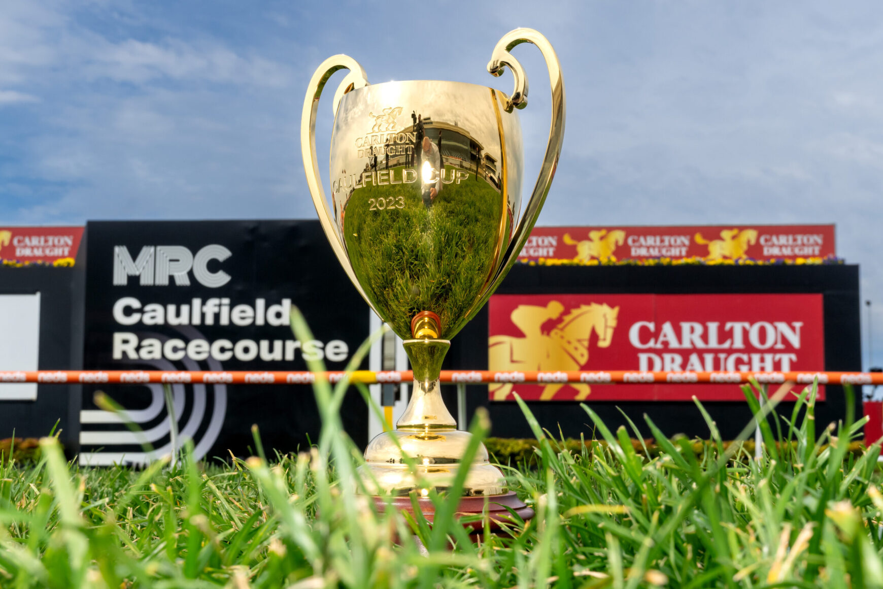 Gold trophy engraved with Carlton Draught Caulfield Cup 2023, standing in grass with racecourse signage in the background.