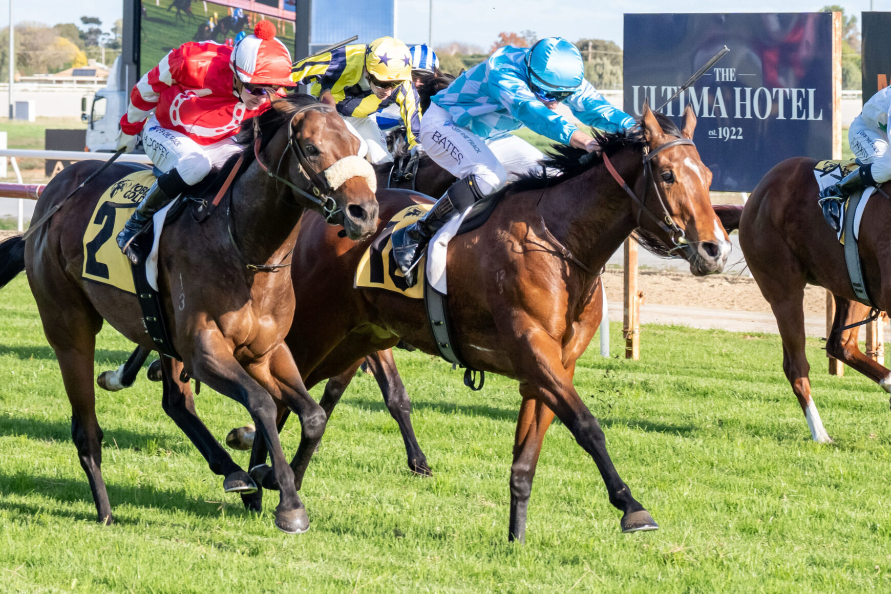 Jockeys in colorful silks ride racing horses on a grassy track, close competition with a large hotel sign in the background reading 'The Ultima Hotel'.