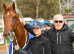 Young woman in a cap and a man in sunglasses stand beside a brown horse at a racetrack, smiling for the camera, with the horse wearing a blue blanket.