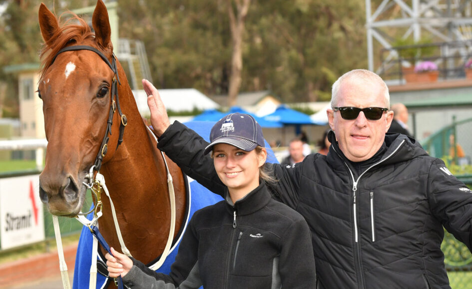 Young woman in a cap and a man in sunglasses stand beside a brown horse at a racetrack, smiling for the camera, with the horse wearing a blue blanket.