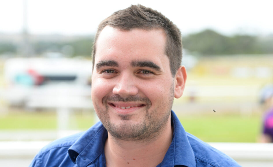 Close-up portrait of a smiling man with short dark hair wearing a blue shirt outdoors.