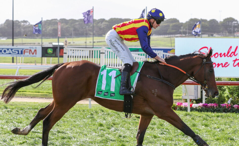 Jockey in red-and-yellow striped silks rides a brown horse with a green number-11 cloth at a racecourse."