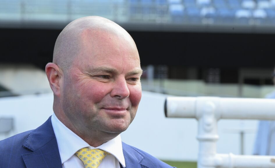 Smiling bald man in a blue suit and yellow patterned tie, outdoors with white piping in the background.