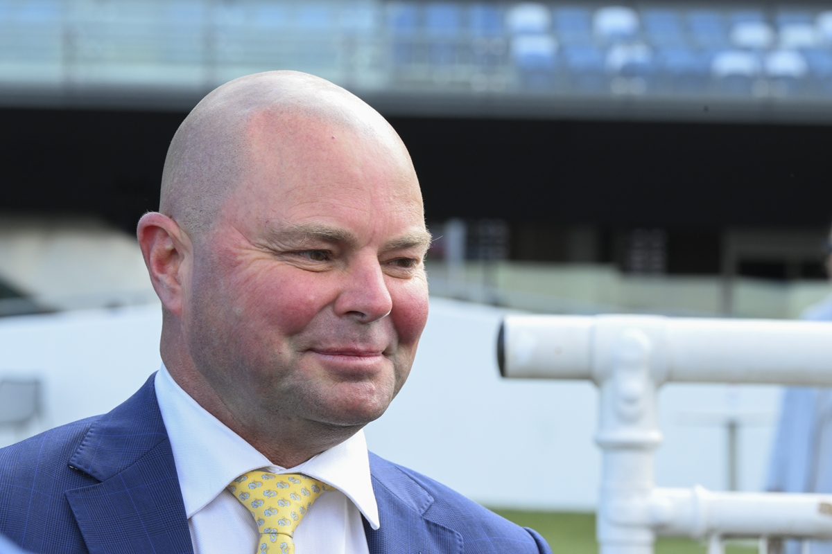 Smiling bald man in a blue suit and yellow patterned tie, outdoors with white piping in the background.