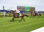 A brown racehorse with a jockey in peach silks leads a photo finish on a green turf track, with a large digital scoreboard in the background reading TAB and spectators off to the side.