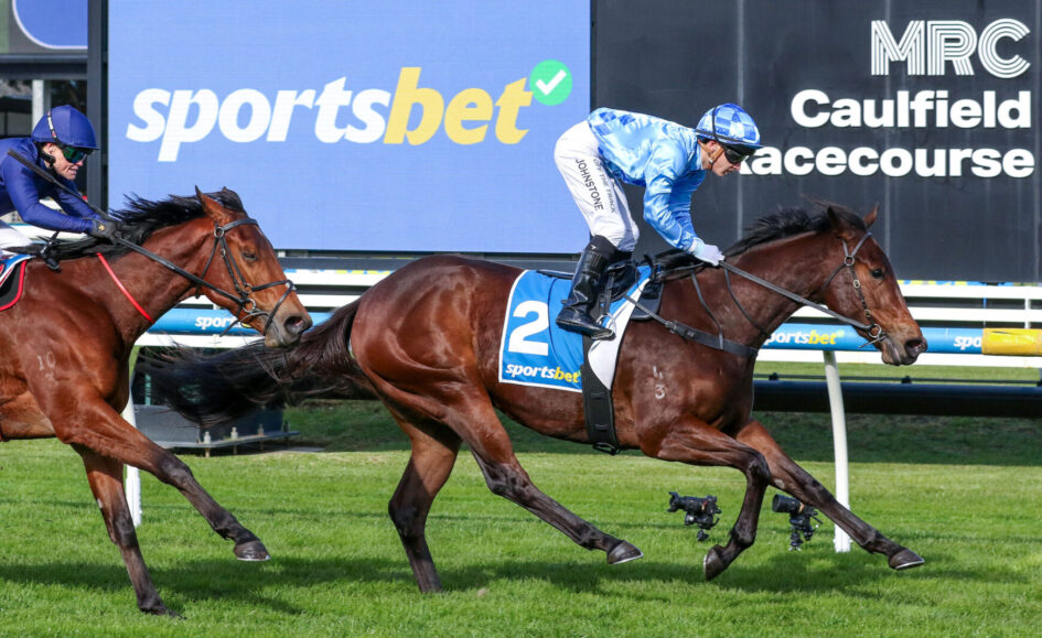 Horse race at Caulfield: front horse in blue silks wearing number 2 saddlecloth, jockey leaning forward as rivals race behind, with 'sportsbet' sign in background.