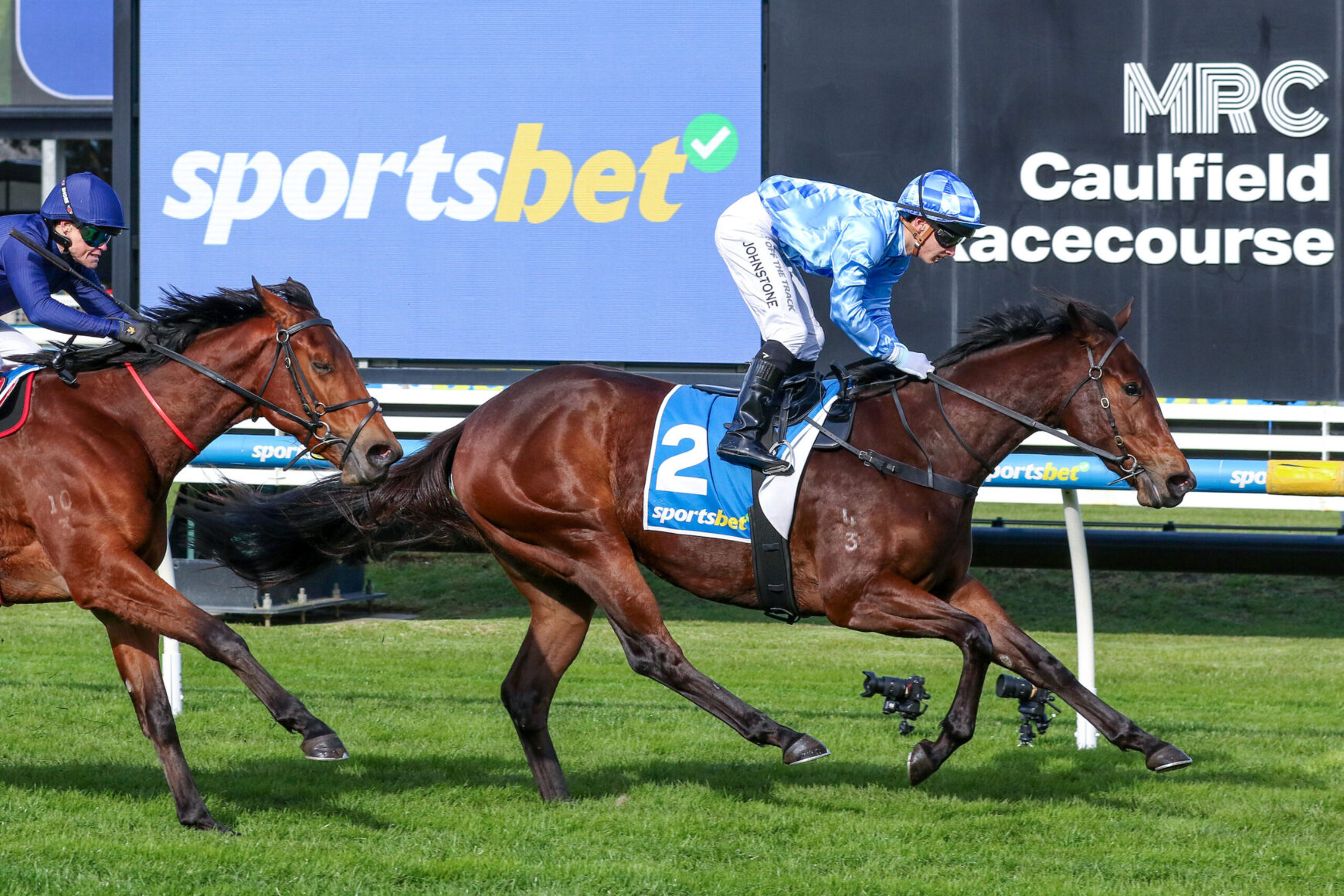 Horse race at Caulfield: front horse in blue silks wearing number 2 saddlecloth, jockey leaning forward as rivals race behind, with 'sportsbet' sign in background.