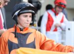 Jockey in orange and black racing silks wearing a helmet, standing at a race venue with handlers in the background.