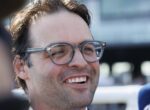 Close-up of a man with gray-framed glasses smiling outdoors, showing stubble on his face