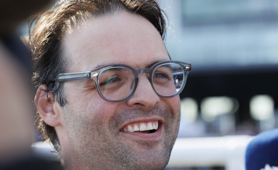 Close-up of a man with gray-framed glasses smiling outdoors, showing stubble on his face