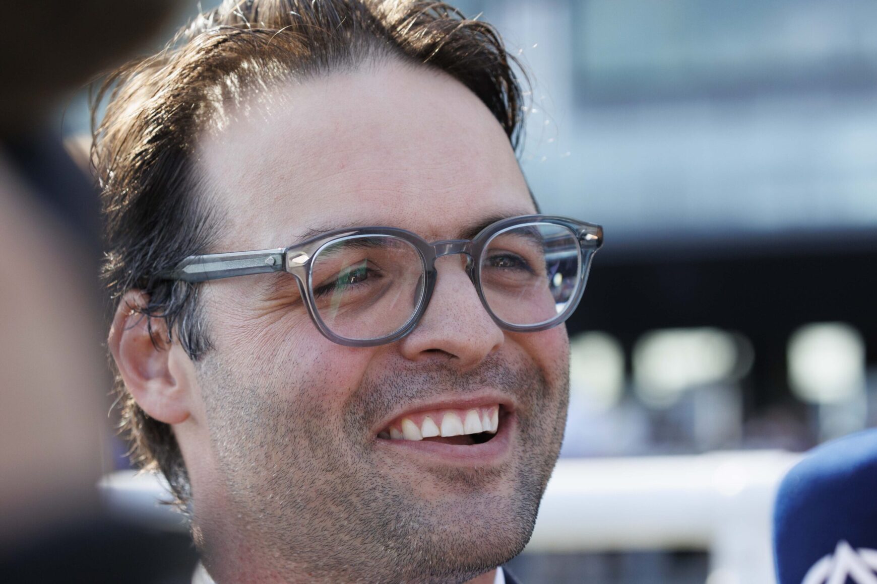 Close-up of a man with gray-framed glasses smiling outdoors, showing stubble on his face