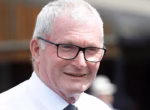 Older man with white hair and black glasses smiling outdoors, wearing a white shirt and tie.