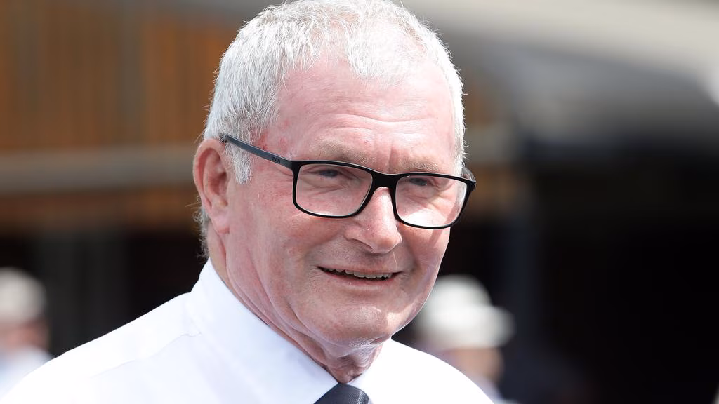 Older man with white hair and black glasses smiling outdoors, wearing a white shirt and tie.