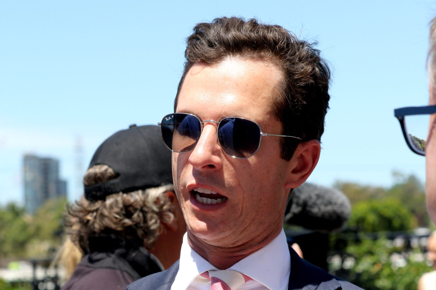 Man in a navy suit and pink tie wearing sunglasses, speaking at an outdoor sunny event.