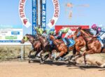 Thoroughbreds and jockeys sprint a dirt racetrack under a blue sky, banners visible in the background.
