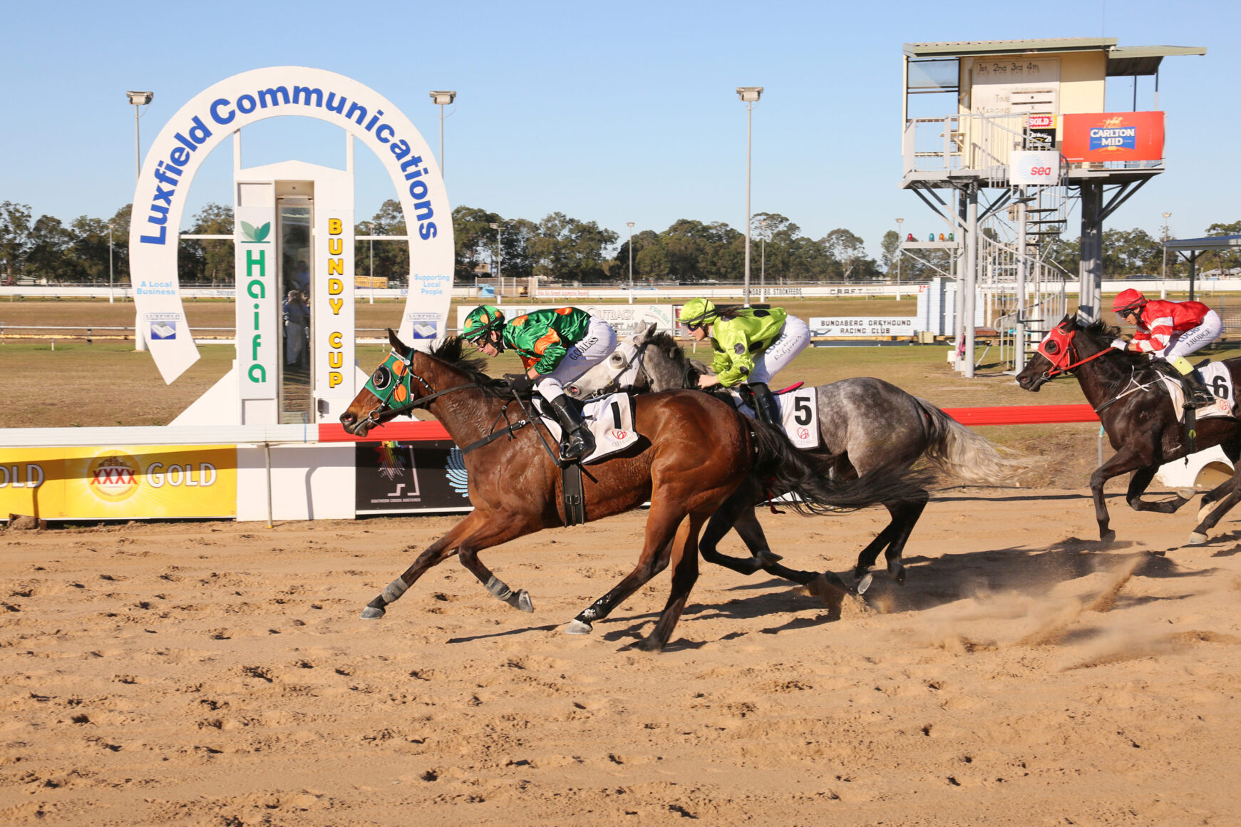 Horse racing at a dirt track with three galloping horses and jockeys in colorful silks, the Luxfield Communications arch in the background.