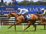 Two jockeys ride thoroughbreds in a close race on a green track, with blue sponsor banners reading Royal Randwick in the background.