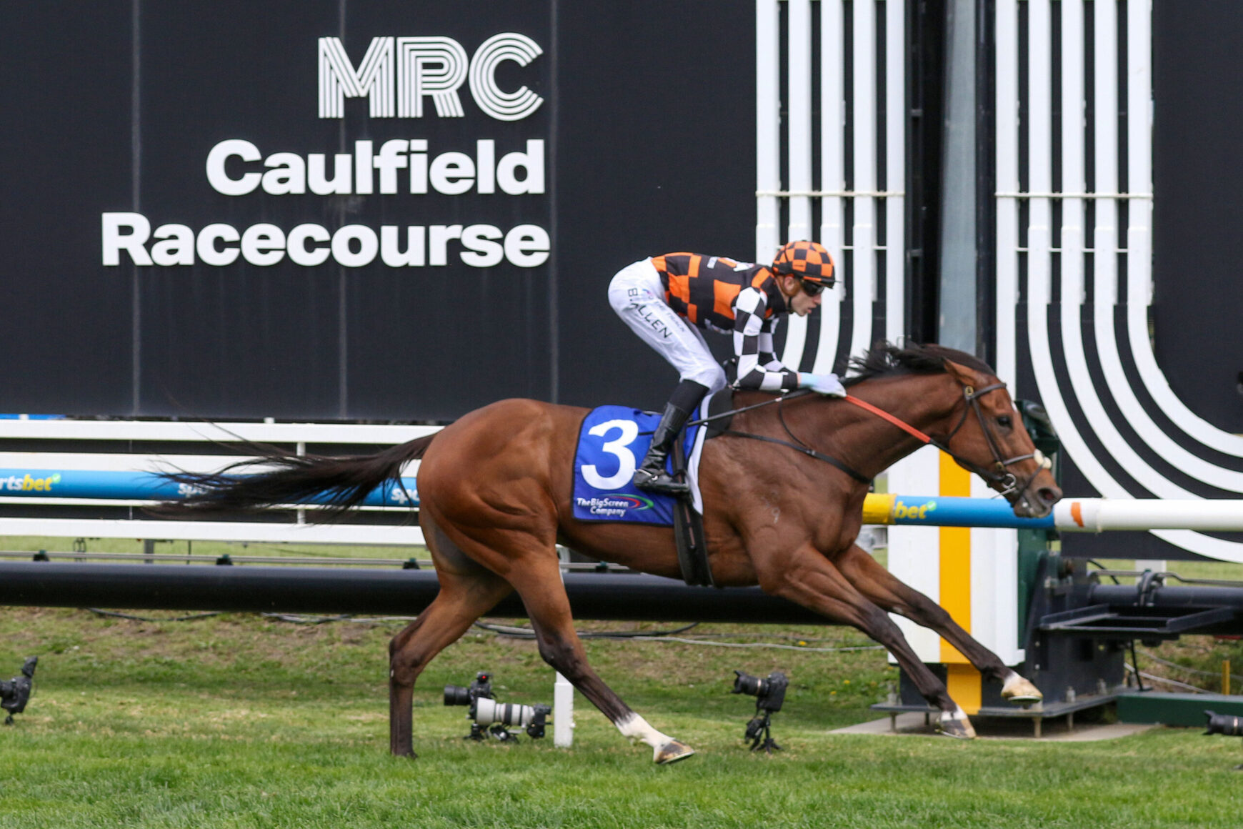 Horse racing scene at Caulfield Racecourse with a jockey in orange-and-black checkered silks riding a bay horse, number 3 saddlecloth