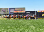 Group of thoroughbred horses with jockeys racing on a green turf track at Caulfield, with large scoreboard and sponsor banners in the background.
