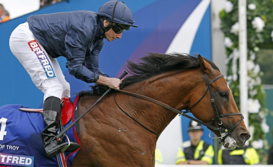Jockey in navy jacket and white pants riding a brown horse during a race, blue barrier in the background, focused forward motion.