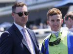 Two men stand together at an outdoor event: one in a navy suit and pink tie wearing sunglasses, the other in high‑visibility racing gear with goggles around his neck.