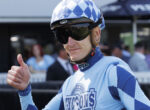 Cyclist in blue kit and a checkered helmet gives a thumbs up at a race event.