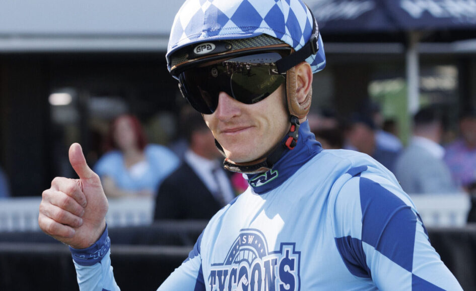 Cyclist in blue kit and a checkered helmet gives a thumbs up at a race event.