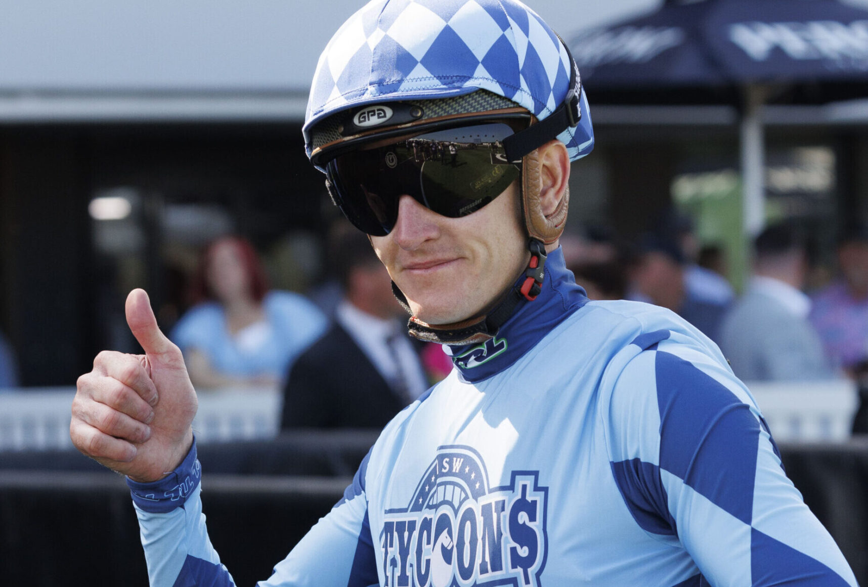 Cyclist in blue kit and a checkered helmet gives a thumbs up at a race event.