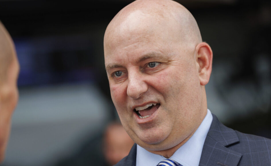 Close-up of a bald man in a blue suit speaking with his mouth open.