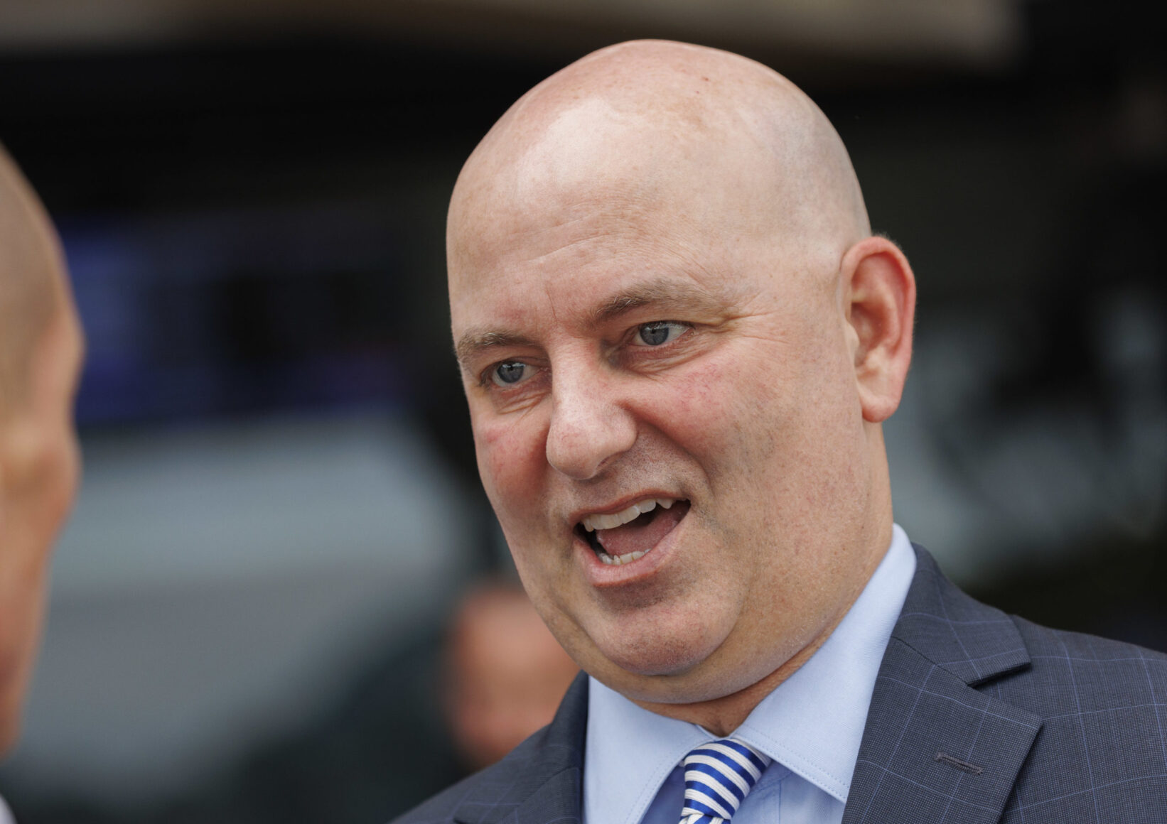 Close-up of a bald man in a blue suit speaking with his mouth open.