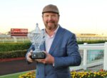 Man in a blue suit holds a crystal trophy at a racetrack, smiling at the camera with a cap on, sunset in the background