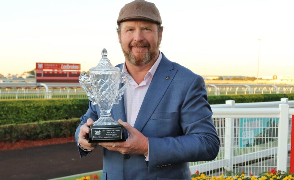 Man in a blue suit holds a crystal trophy at a racetrack, smiling at the camera with a cap on, sunset in the background