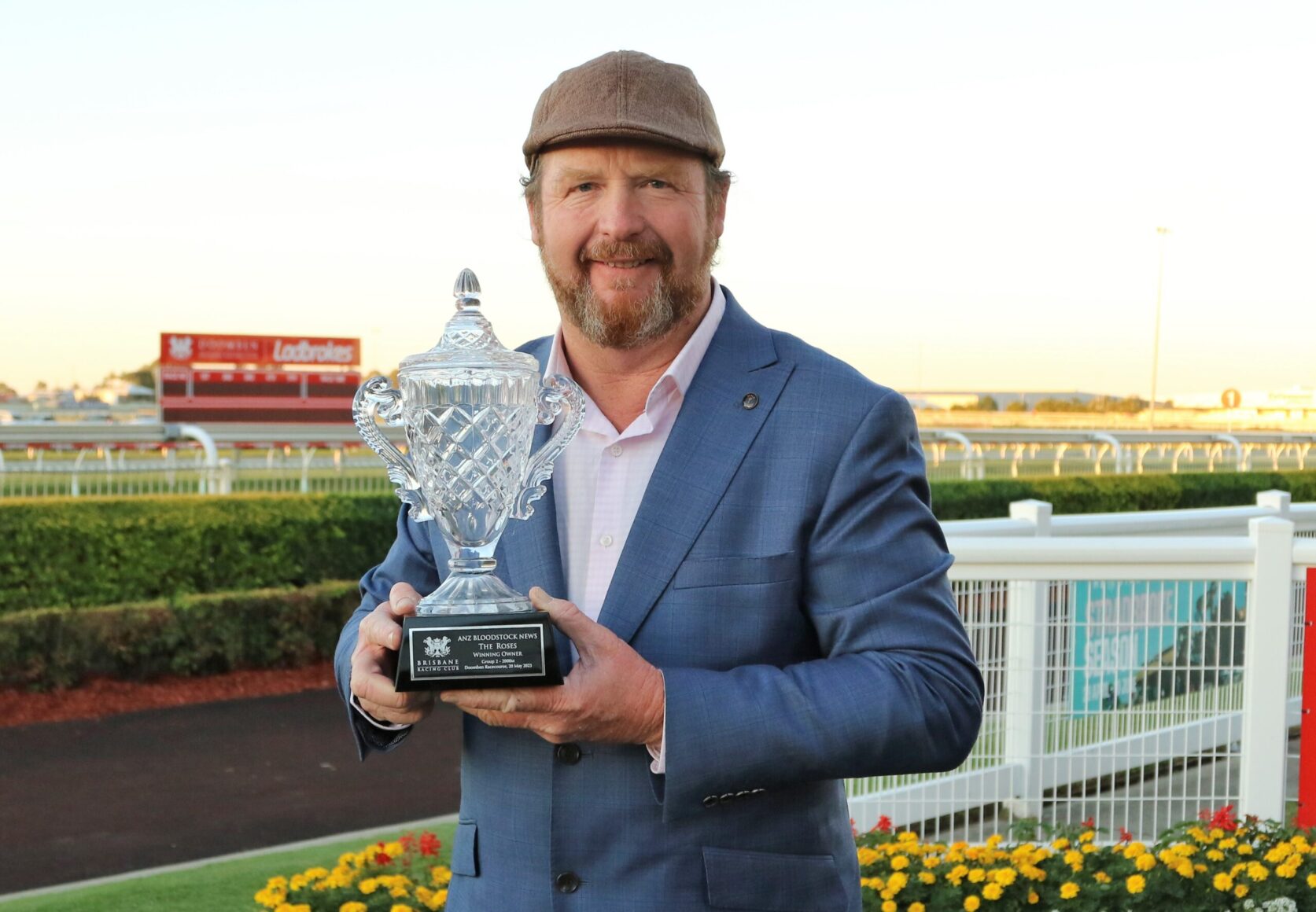 Man in a blue suit holds a crystal trophy at a racetrack, smiling at the camera with a cap on, sunset in the background