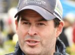 Close-up of a man wearing a dark baseball cap and quilted jacket, speaking outdoors with a blurred background.