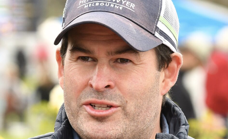 Close-up of a man wearing a dark baseball cap and quilted jacket, speaking outdoors with a blurred background.