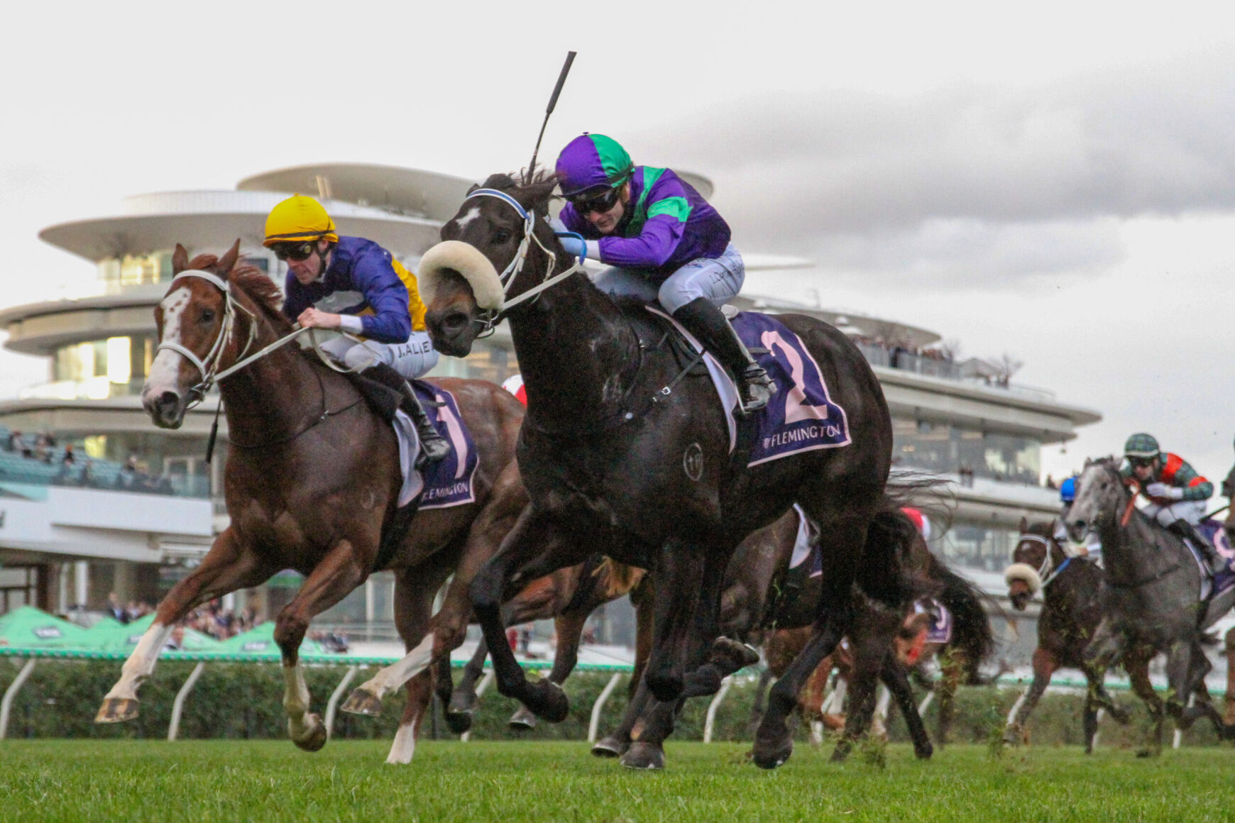 Three jockeys in colorful silks ride sprinting horses mid-jump during a race at Flemington, with a grandstand in the background.